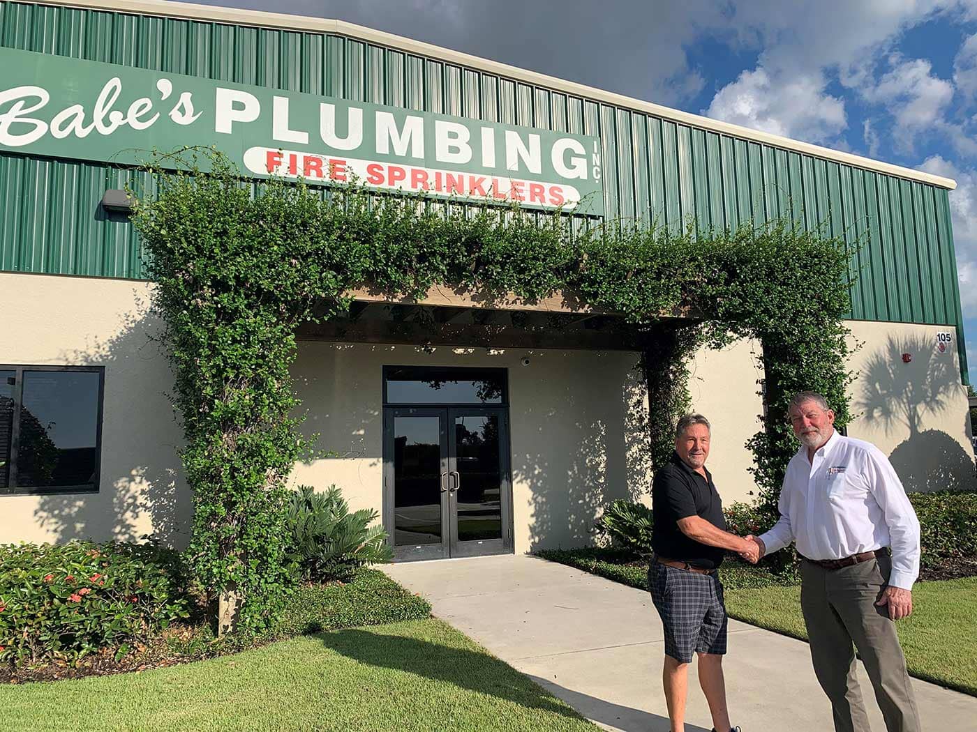 Two men shaking hands in front of a building with signage that reads "Babe’s Plumbing Fire Sprinklers.