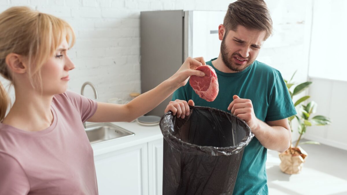 A woman throws food into the garbage disposal as a man holding a trash bin looks at it with a grimace, troubled by the disposal only humming.