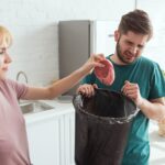 A woman throws food into the garbage disposal as a man holding a trash bin looks at it with a grimace, troubled by the disposal only humming.