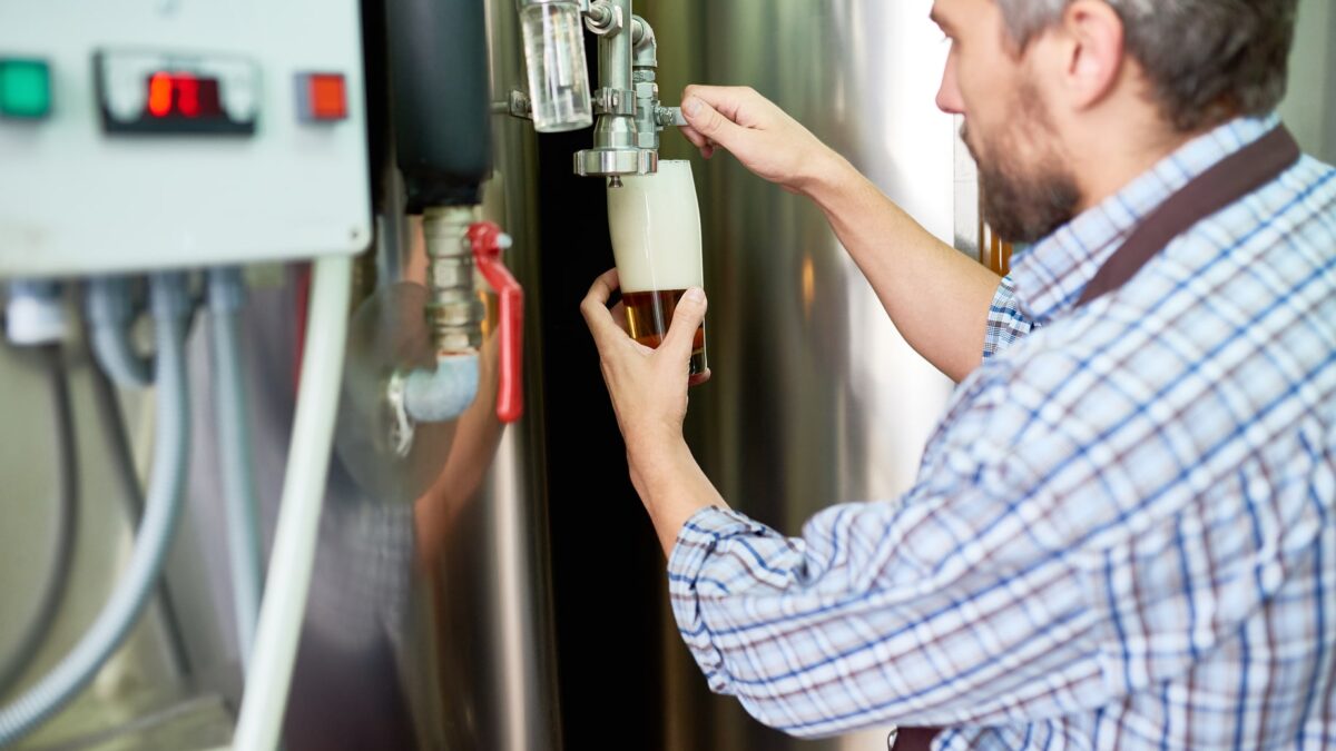 Man collecting a sample from a steel tank at a brewery or factory to monitor clogged water heater drainage.